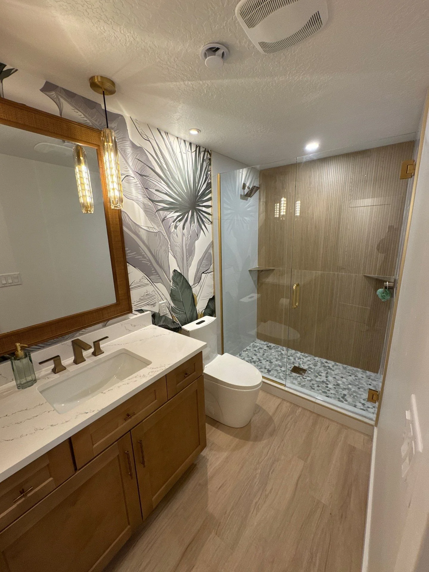 Custom bathroom with framed mirror, gold pendants, glass-enclosed shower, and tropical wallpaper