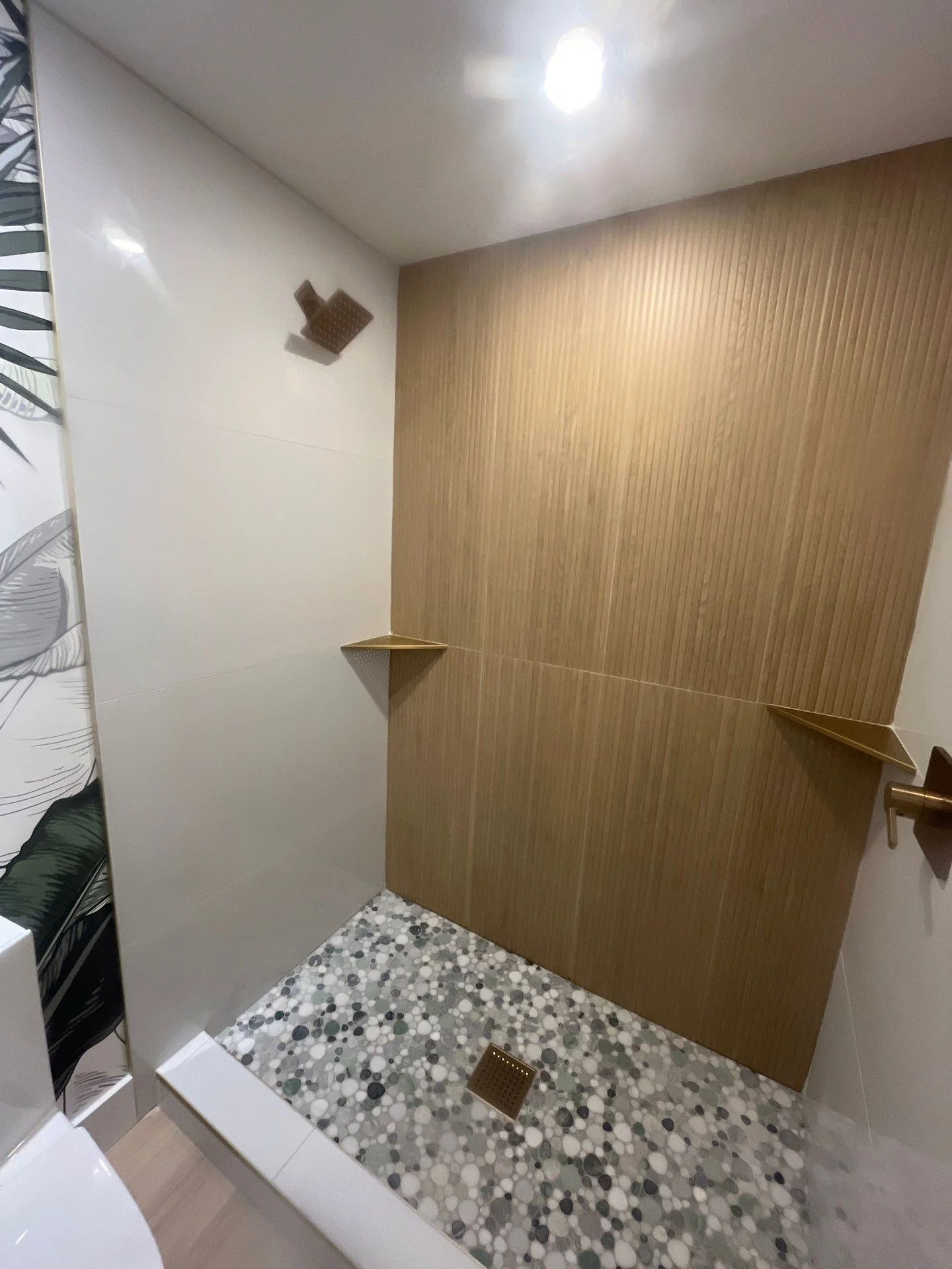 Walk-in shower with wood-look slat tile back wall, gold corner shelves, and pebble mosaic floor